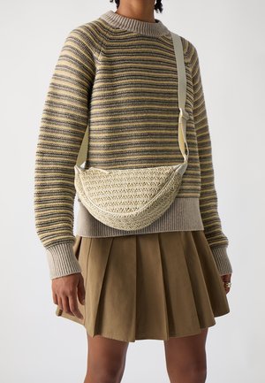 Personne portant un pull beige à rayures et une jupe plissée marron, portant un sac bandoulière tissé de couleur crème avec une sangle ajustable.