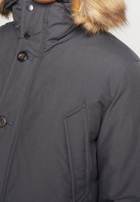 Manteau d'hiver gris foncé avec capuche bordée de fourrure, fermetures à boutons et pressions, et poche latérale verticale au niveau de la poitrine.