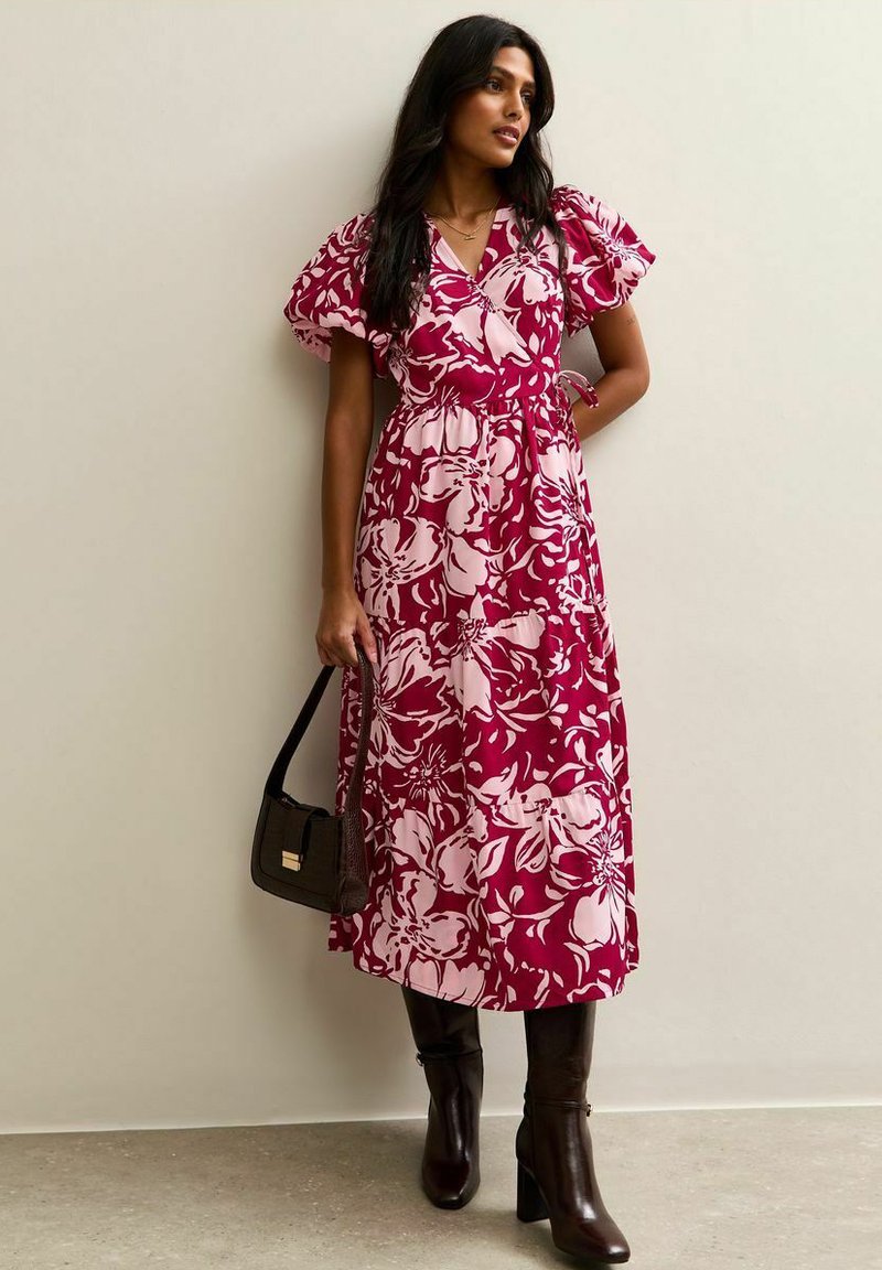 Robe maxi à imprimé floral en bordeaux et rose pâle, avec manches bouffantes et taille nouée. Associée à des bottes hautes marron et un sac sombre.