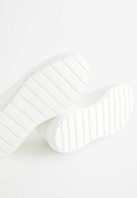 Baskets blanches avec une semelle en caoutchouc texturée présentant des rainures horizontales, conçues pour l'adhérence et la durabilité. Bout rond et design minimaliste.