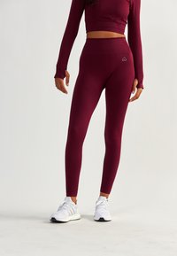 Bordowe legginsy z wysokim stanem, prążkowaną teksturą i logo na biodrze, w połączeniu z białymi butami sportowymi.