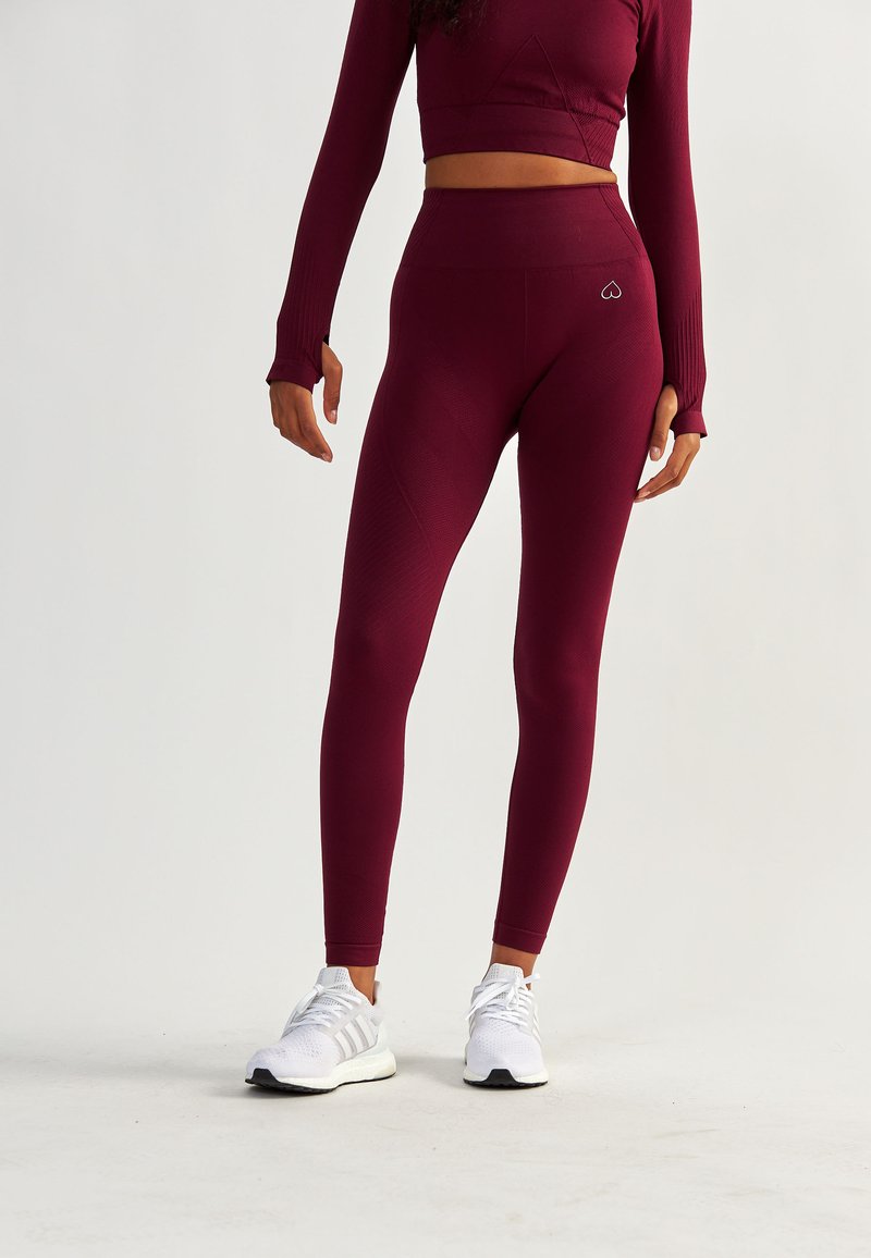 Bordowe legginsy z wysokim stanem, prążkowaną teksturą i logo na biodrze, w połączeniu z białymi butami sportowymi.