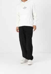 Witte hoodie met een voorzak en een rood-blauw logo, gecombineerd met zwarte broek en witte sneakers. Eenvoudige, casual outfit getoond.