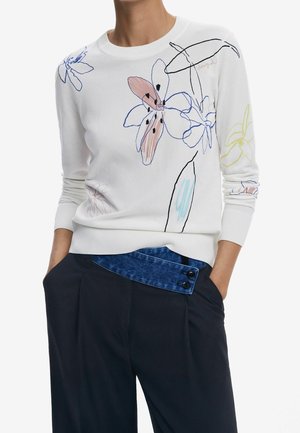 Pull blanc à manches longues avec des dessins floraux abstraits au trait en bleu, rose, noir et jaune, associé à un pantalon bleu marine avec un détail asymétrique en denim à la taille.