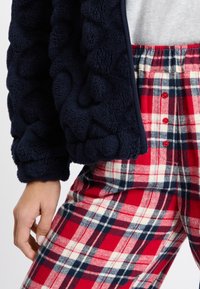 Marineblaues Fleece-Jackett mit strukturiertem Oberflächen, mit Reißverschluss, kombiniert mit rot-weißen und marineblauen karierten Hosen mit Knöpfen und elastischem Bund.