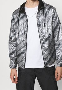 Silver vindjacka med diagonala svarta ränder och huva. Har en framdragkedja och elastiska ärmslut. Bärs över en vit t-shirt.