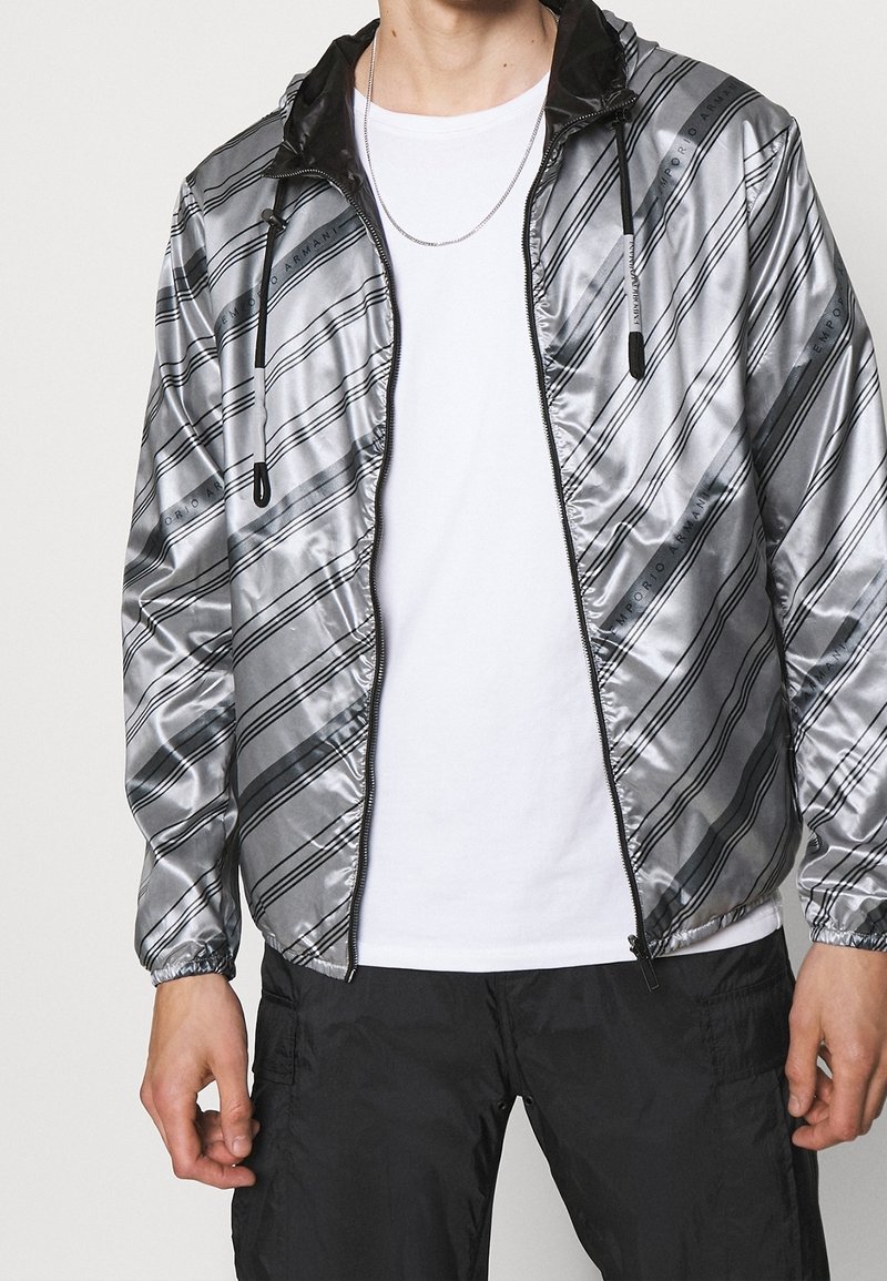 Silver vindjacka med diagonala svarta ränder och huva. Har en framdragkedja och elastiska ärmslut. Bärs över en vit t-shirt.