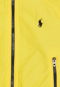 Žlutá zipová bunda vyrobená z hladké tkaniny, se černým zipem, černým vyšitým logem a značkovým zipovým táhlem.