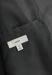 Tejido negro con una textura suave, con una etiqueta cosida que dice "NEXT", talla "M" y "FABRICADO EN CHINA". Detalle sutil de un bolsillo visible.