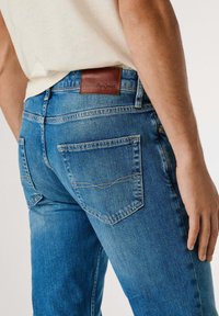 Niebieskie dżinsy o dopasowanym kroju, z tylnymi kieszeniami i brązową skórzaną metką przy pasku. Materiał ma fakturowane wykończenie.
