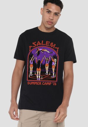 Young man wearing a black T-shirt featuring five cheerleaders in a pentagram circle levitating a sixth girl at night, titled "Salem Summer Camp '78."