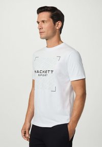 Bílá bavlněná tričko s centrálním černým logem 'HACKETT SPORT', rámované geometrickými akcenty s jemným vzorem. Krátké rukávy, kulatý výstřih.