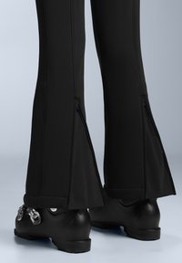 Černé kalhoty s rozšířenými nohavicemi, které mají boční zipy. Materiál vypadá hladce a jsou doplněny lesklými černými botami.