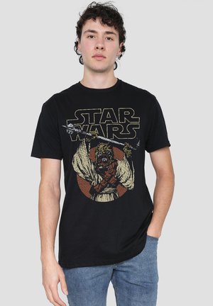 Junger Mann trägt ein schwarzes Star Wars T-Shirt mit einem Tusken Raider, der eine Waffe hält, kombiniert mit blauen Jeans vor schlichtem Hintergrund.