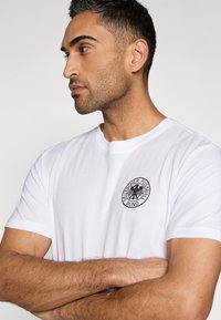 Λευκό βαμβακερό T-shirt με στρογγυλή λαιμόκοψη, κοντά μανίκια και μαύρο κυκλικό λογότυπο με αετό και κείμενο στο στήθος.