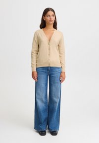 Cardigan beige à col en V avec fermeture à bouton et manches longues, associé à un jean bleu à jambes larges. Texture lisse, coupe décontractée et design simple.