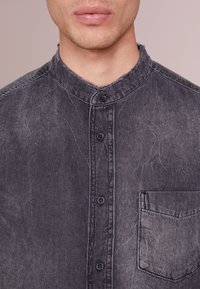 Primer plano de una persona que lleva una camisa de mezclilla negra desvaída, con botones y un cuello chino, y un bolsillo en el lado derecho del pecho.