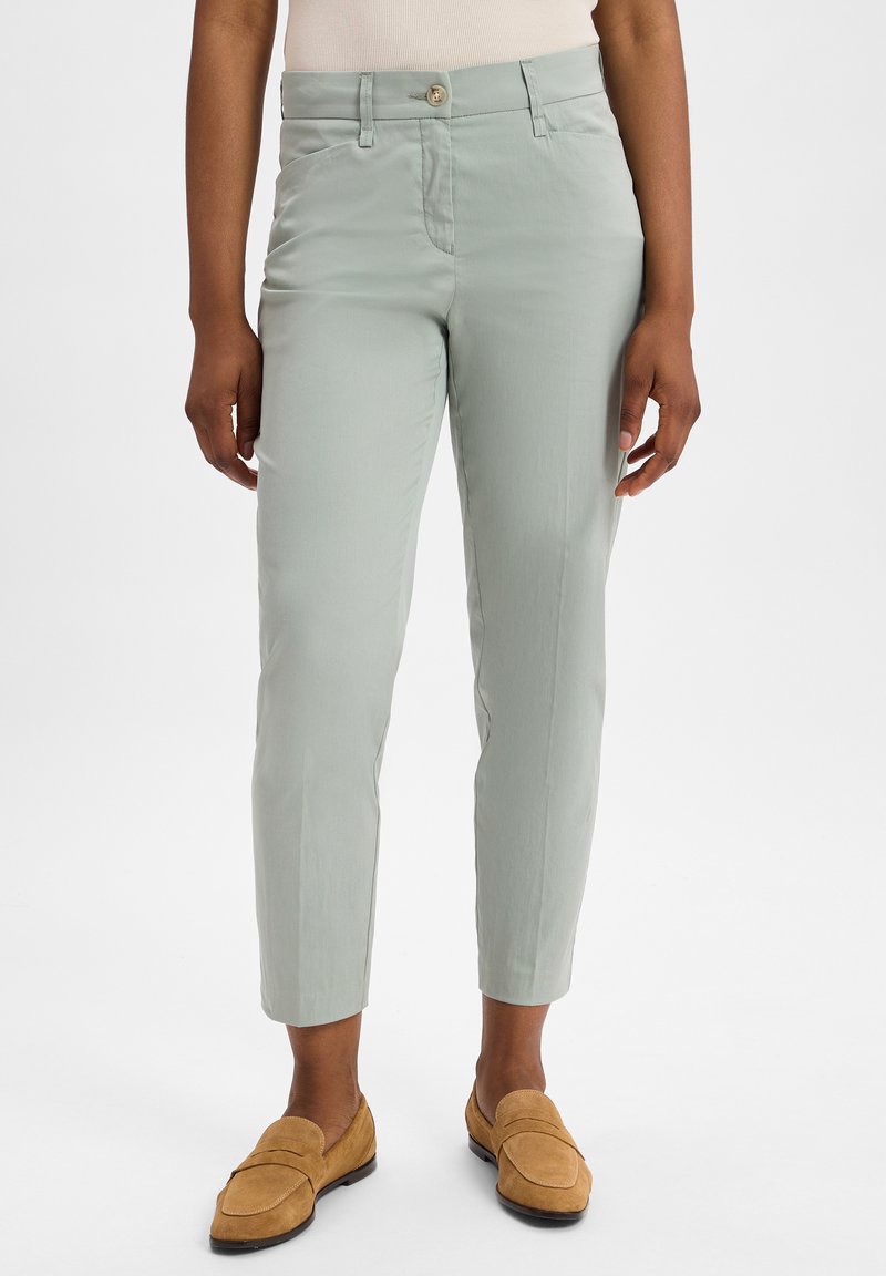 Hellgrüne, schlank geschnittene Hose mit glatter Textur, ausgestattet mit einem frontalen Knopfverschluss und zwei seitlichen Taschen, kombiniert mit hellbraunen Mokassins.