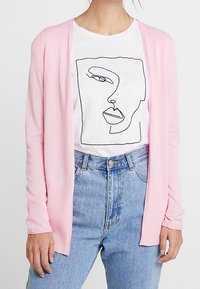 Cardigan long rose pâle en tricot doux, design ouvert devant. Porté sur un t-shirt blanc avec un dessin au trait noir d'un visage et un jean taille haute bleu.
