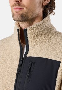 Beige fleece jacket with high collar and black nylon accents. Features a front zipper and logo detailing on the chest pocket.