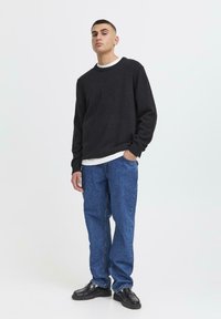 Homme debout les mains dans les poches, portant un pull noir, un t-shirt blanc, un jean bleu et des mocassins noirs devant un fond uni.