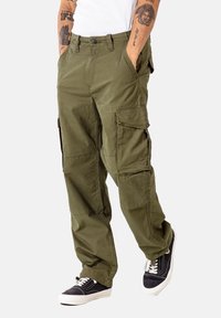 Olivengrüne Cargo-Hosen mit mehreren Taschen, einem Knopfverschluss und einer lockeren Passform, kombiniert mit schwarzen Sneakers und einem weißen Hemd.