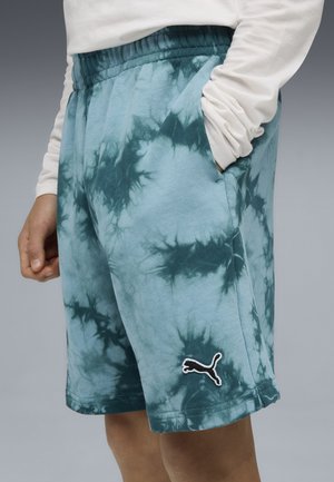 Persoon draagt blauw tie-dye short met elastische tailleband en Puma-logo op het linkerbeen, één hand in de zak, witte longsleeve zichtbaar.