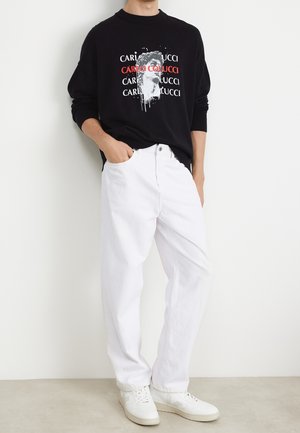 Black sweatshirt with a graphic print in red and white, paired with white denim jeans and white sneakers, standing against a plain background.