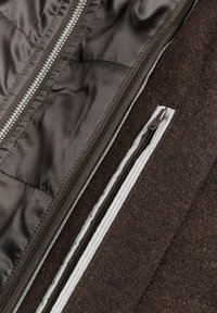 Close-up van bruine jasstof met twee parallelle donkere ritsen, één met witte biezen langs de naad.