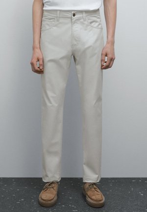 Person wearing light beige pants and brown shoes, standing against a simple gray background. Hands are relaxed at the sides.