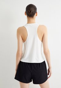 Top deportivo blanco de ajuste relajado, con diseño de espalda nadadora y textura suave, combinado con pantalones cortos negros. Estilo limpio y minimalista.