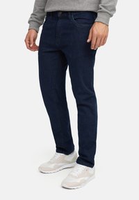 Homme portant un jean slim bleu foncé et des baskets blanches avec des détails gris clair et beige, debout devant un fond blanc.