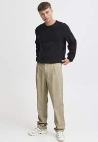 Schwarzer Strickpullover mit Rundhalsausschnitt, kombiniert mit hellbeigen Hosen. Das Outfit wird durch weiße und graue Turnschuhe komplettiert.