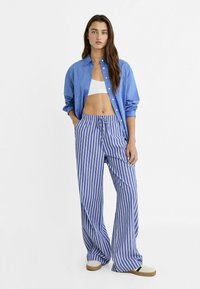 Kvinna stående med händerna i fickorna, klädd i blå skjorta med knappar, vit crop top, blå och vita randiga vida byxor samt vita sneakers.