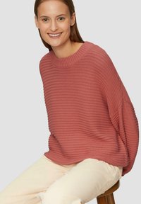 Roze geribbelde gebreide trui met een brede halslijn en een losse pasvorm, gecombineerd met crèmekleurige broek, zittend op een kruk.