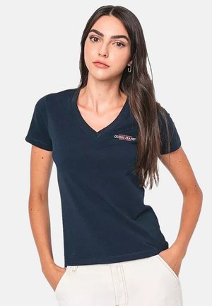 Jeune femme aux longs cheveux bruns portant un T-shirt bleu marine à col en V avec le logo "Guess Jeans", mains dans les poches de son pantalon blanc, expression neutre.