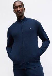 Sweatshirt navy cu fermoar din țesătură texturată, cu manșete și talie ribbed. Prezentă un logo brodat pe piept și buzunare laterale.