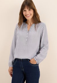 Blusa a maniche lunghe con bottoni, a righe bianche e navy, con scollo a V e vestibilità rilassata, abbinata a jeans scuri.
