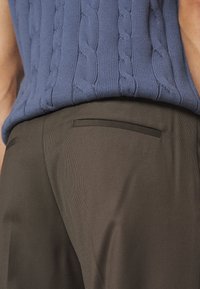 Gros plan sur une personne portant un pull bleu en maille torsadée rentré dans un pantalon marron taille haute avec une poche passepoilée à l'arrière.