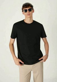 Schwarzes T-Shirt mit kurzen Ärmeln aus leichtem Stoff, mit rundem Ausschnitt und geradem Schnitt, kombiniert mit beigen Hosen, getragen von einem Modell.