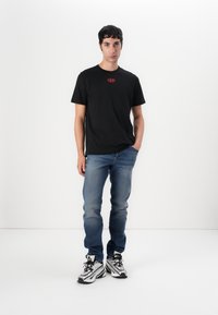 Zwarte katoenen t-shirt met een rode logo op de borst, gecombineerd met blauwe spijkerbroeken en zwart-witte sneakers met textuurdetails.