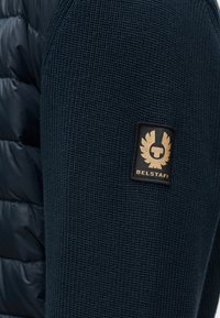 Σκούρο ναυτικό πλεκτό ύφασμα με ραβδωτή υφή· διαθέτει μαύρη ετικέτα με χρυσές λεπτομέρειες και το λογότυπο "Belstaff" στο μανίκι.