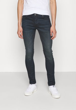 Vaqueros ajustados de lavado oscuro con una textura suave, que cuentan con un diseño clásico de cinco bolsillos y cierre de cremallera frontal. Combinados con zapatillas blancas.