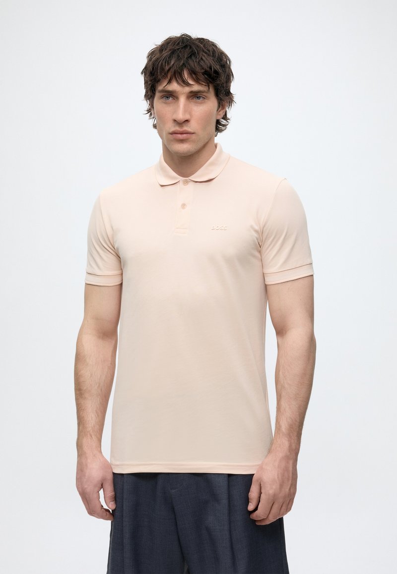 Homme aux cheveux courts et foncés, portant un polo beige clair à manches courtes et un pantalon foncé, debout sur un fond uni.