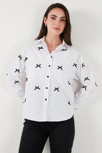 Vrouw draagt een wit overhemd met zwarte X-vormige patronen en zwarte broek, staand tegen een effen witte achtergrond.