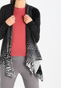 Svart och gråmönstrad cardigan med draperad front, blandade djurmönster, mjuk textur och långa ärmar över en röd topp och grå byxor.