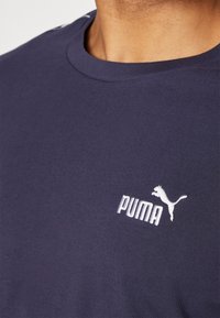Temno modra majica iz bombažnega mešanega materiala, z okroglo ovratnico in belim vezenim logotipom Puma na prsih. Gladka teksturna površina.