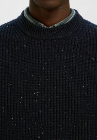 Tamsiai mėlynas megztinis su raštu, aukštu kaklu ir daugybe spalvotų dėmių. Dėvimas ant langelių raštuoto marškinio.