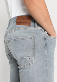 Jeans de mezclilla azul claro con parche de cuero marrón etiquetado "TOMMY HILFIGER" en la cintura, con costuras naranjas y bolsillos laterales.