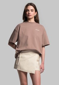 Brun oversized t-shirt med korte ærmer og rund hals, med hvid logo tekst. Sammen med en lys beige omslagsskjørt med bindebånd.